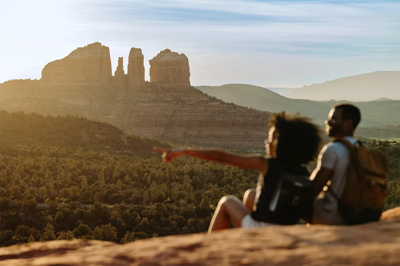 A man and a woman sit on red rock formations at sunset admiring the view