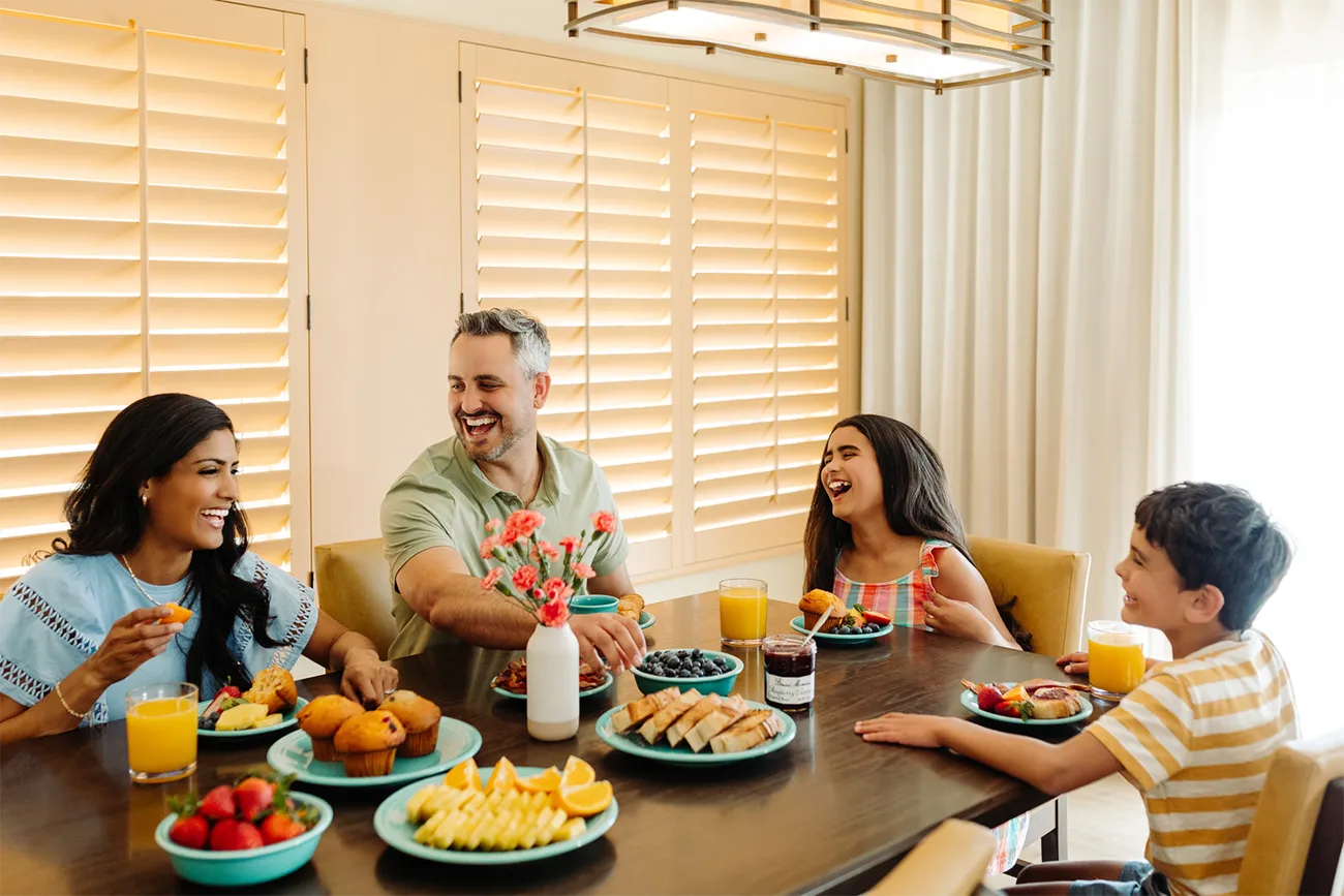 A man, woman, girl and boy sit around a dining room table sharing a meal in a villa room