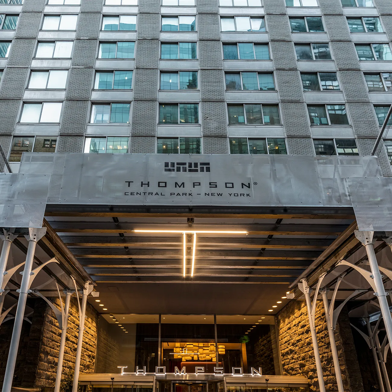The entrance of Thompson Central Park, a modern high-rise building with a glass and steel facade, illuminated with warm lighting