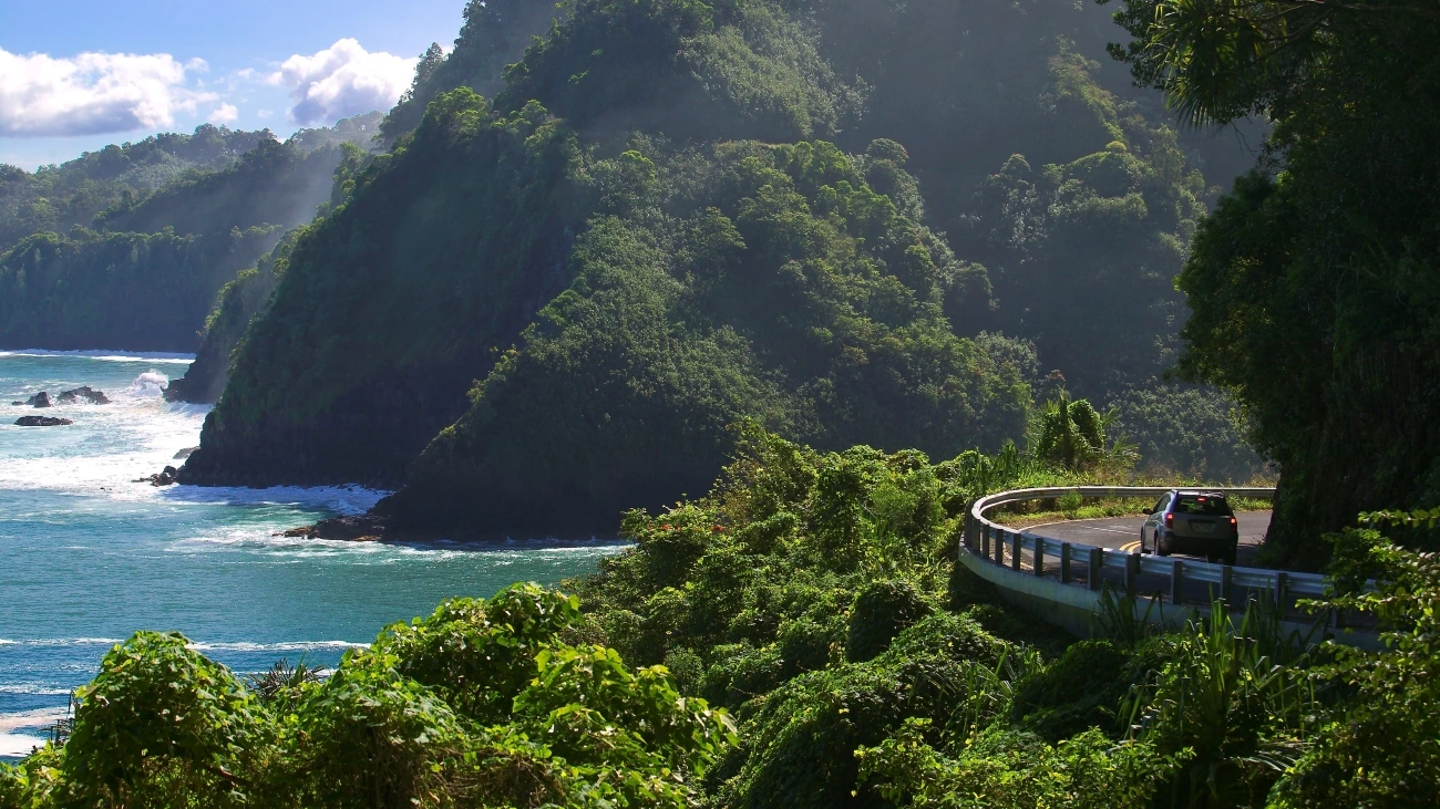 A scenic winding seaside road in Hawaii, with waves breaking along a lush, mountainous coast