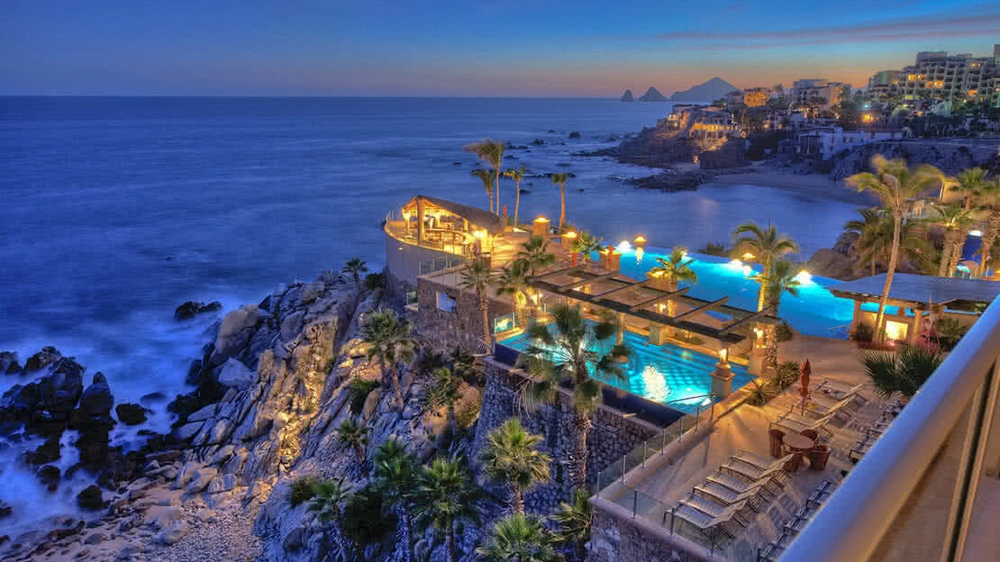 The sun sets over the ocean while an infinity pool and outdoor pool area light up