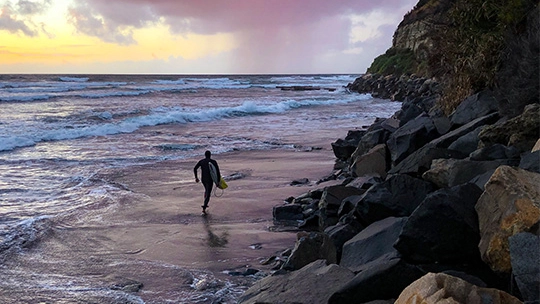 Surfer with surfboard walking along a rocky beach at dawn, ready to catch his next wave
